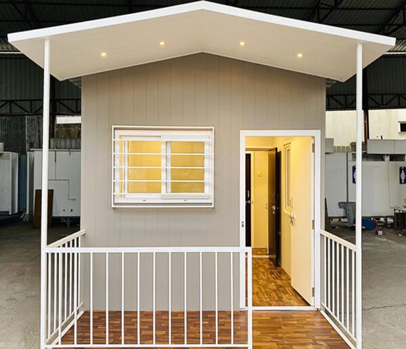 A modern prefabricated cabin with a white slanted roof, beige walls, and a small porch enclosed by white railings. 