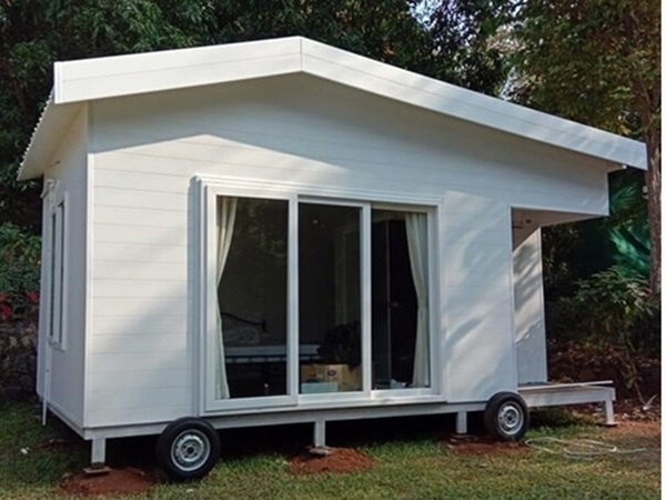 A white mobile cabin with a slanted roof, large sliding glass doors, and wheels, placed on grassy ground with trees in the background.